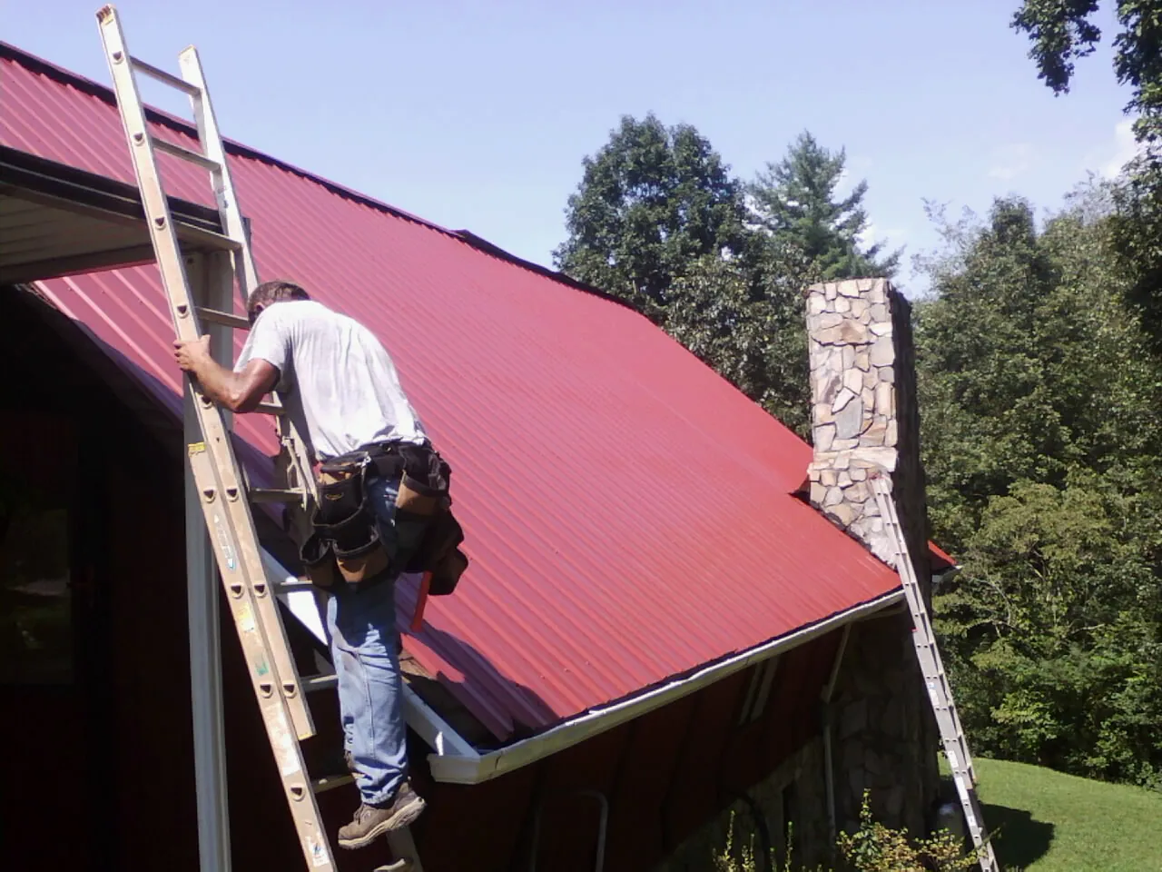 Roofer on a ladder beside a red metal roof and stone chimney — Soffit & Fascia Repair by Roofing Brothers in Mounds